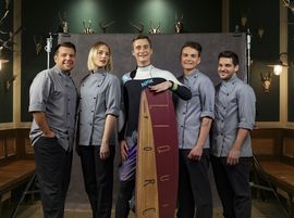 In der Mitte befindet sich der Küchenleiter des Restaurants im Neoprenanzug mit Surfbrett und rechts und links von ihm stehen insgesamt vier weitere Mitarbeiterinnen und Mitarbeiter des Küchenteams in  grauen Kochjacken lachend nebeneinander.