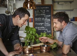 Zwei Mitarbeiter der Küche stehen sich in Kochjacken in der Küche gegenüber und blicken zueinander. Mittig im Bildhintergrund hängt die aktuelle Speisekarte des Restaurants Pfistermühle an der Wand.