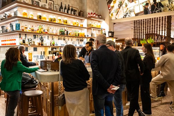 Eine Gruppe von Menschen steht und sitzt an einer gut beleuchteten Hotelbar, während der Barkeeper Getränke zubereitet
