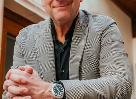 Mann mit Brille im karierten Sakko vor Hotelfassade, freundlich blickend