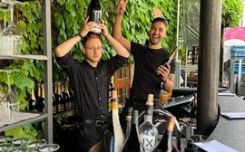 Zwei Barkeeper scherzen hinter Outdoor-Bar mit Weinflaschen und Reben