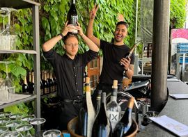 Zwei Barkeeper scherzen hinter Outdoor-Bar mit Weinflaschen und Reben