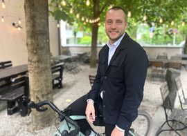 Mann im Anzug auf Fahrrad im Biergarten mit Lichterkette und Kiesboden